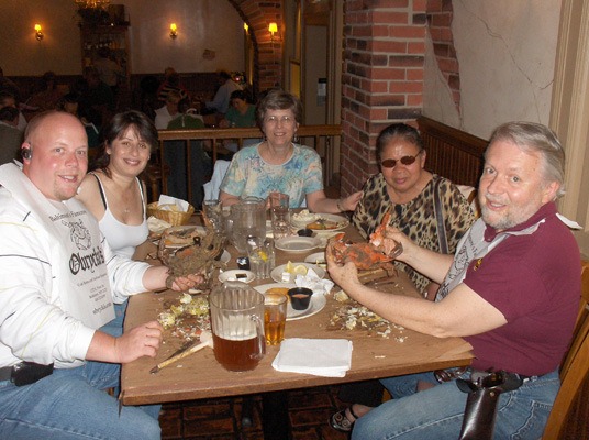 Seated around a table, which is coverd with brown paper, are Paul, Jomania, Dona, Nanta and Fred.  On the table is a pitcher of beer and lots of empty crab shells. Fred and Paul are holding up crabs (they are about a foot wide, from the end of one pincher to the other).